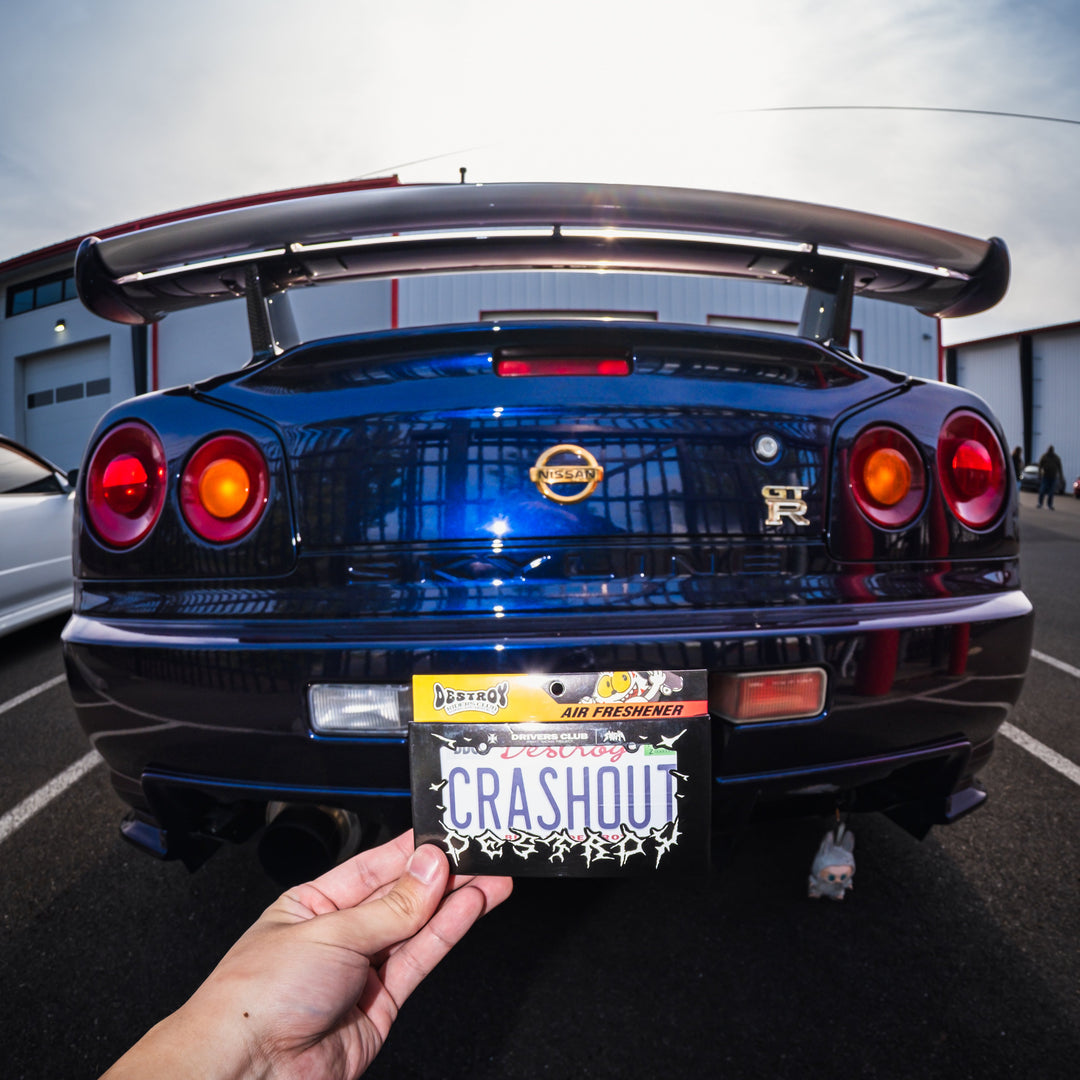 Person holding a 'CRASHOUT' license plate in front of a blue car with a visible license plate.
