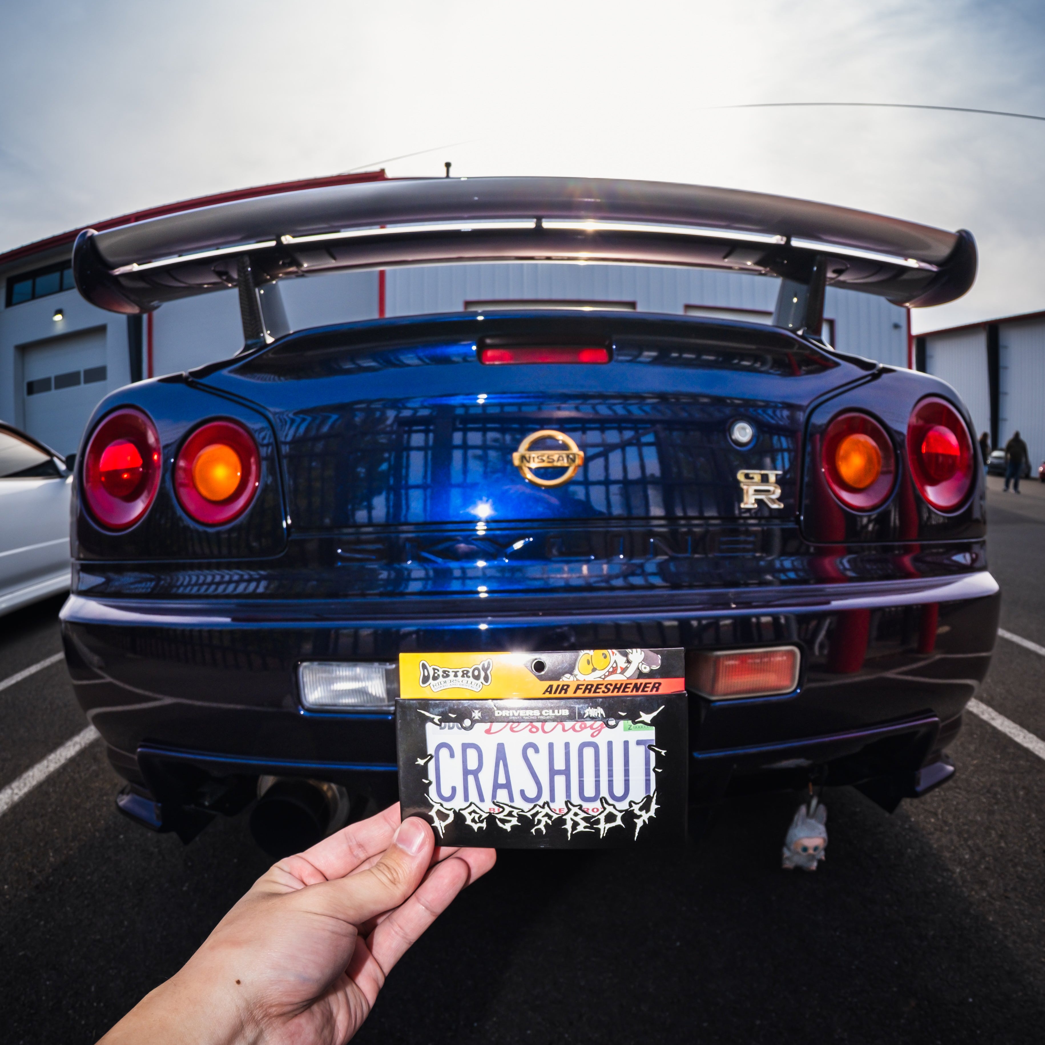 Person holding a 'CRASHOUT' license plate in front of a blue car with a visible license plate.