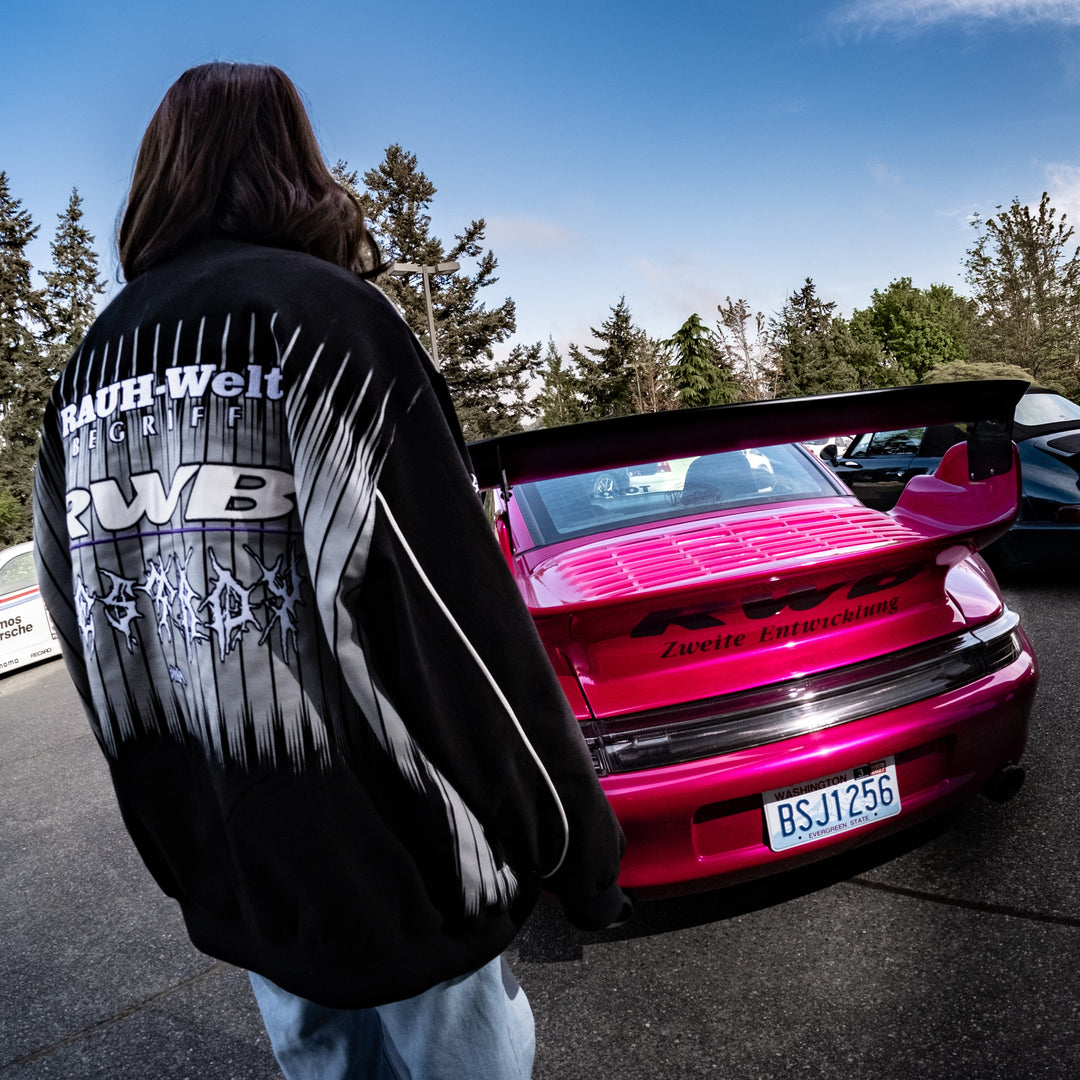 Person wearing a black jacket with white text standing next to a pink car on a sunny day.