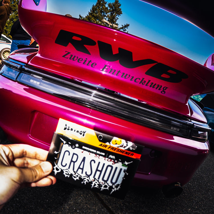Pink car with RWB badge and 'CRASHOUT' license plate held by a hand.