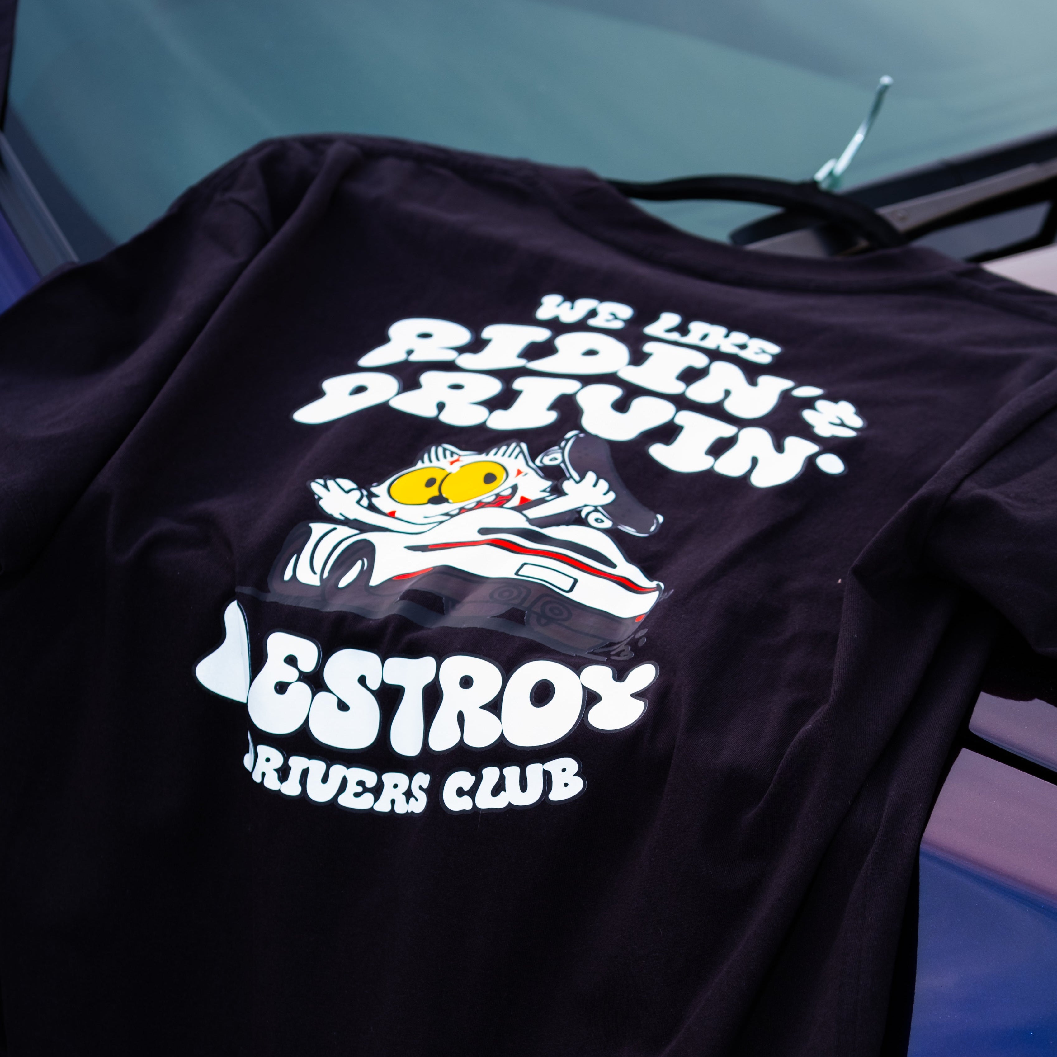 Black t-shirt with graphic design and text, hanging against a car interior background.