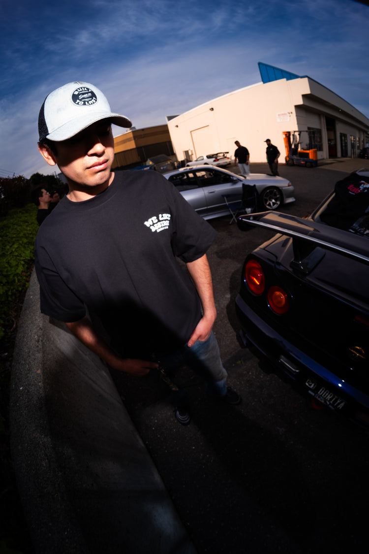 Person wearing a cap and black t-shirt standing next to a car with a building and other people in the background.
