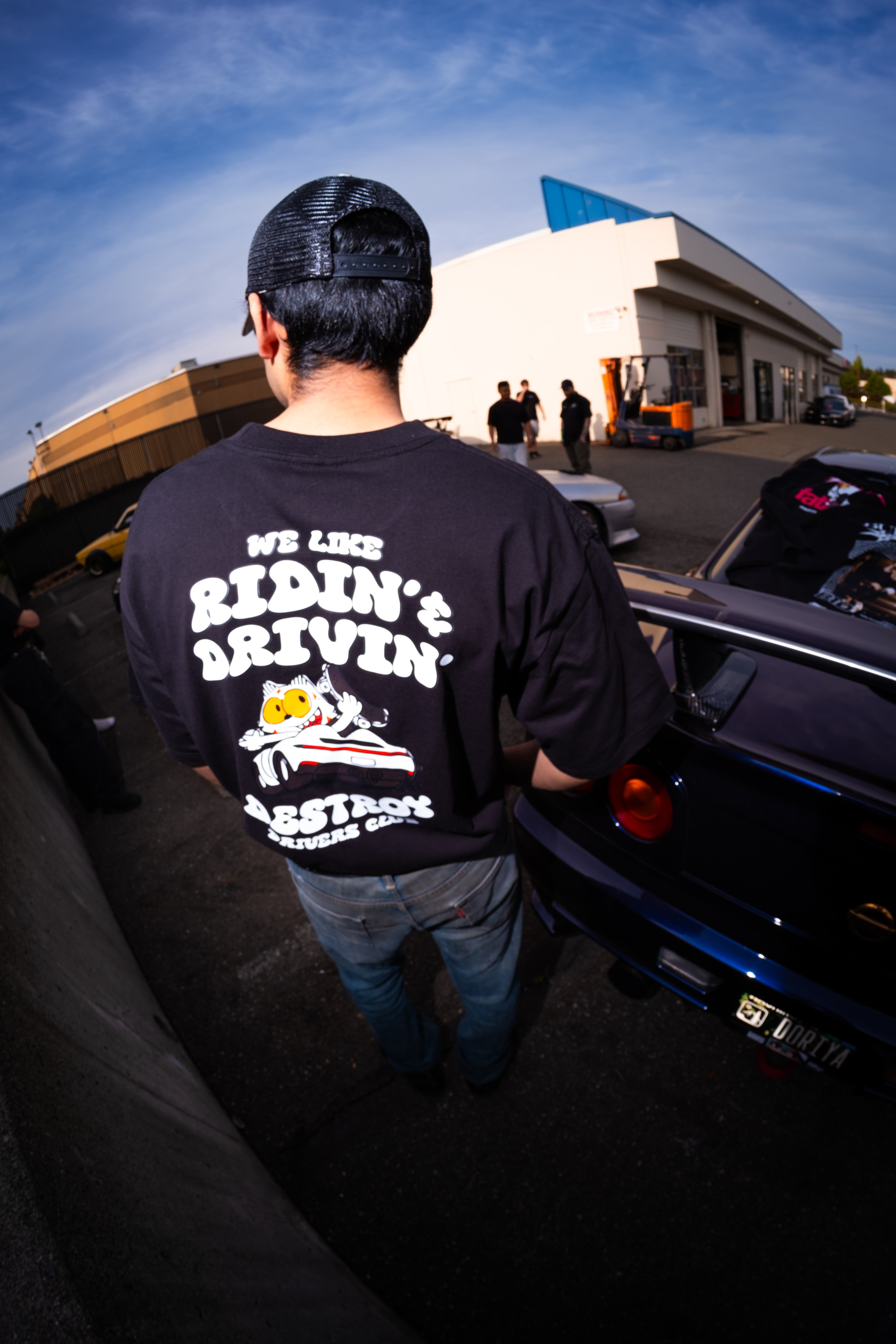 Person wearing a black t-shirt with text and graphics, standing next to a car with an open trunk.