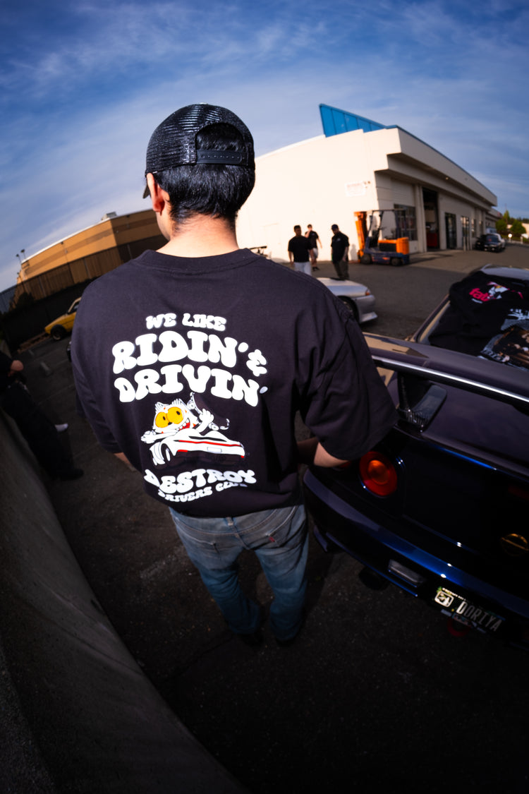 Person wearing a black t-shirt with text and graphics, standing next to a car with an open trunk.