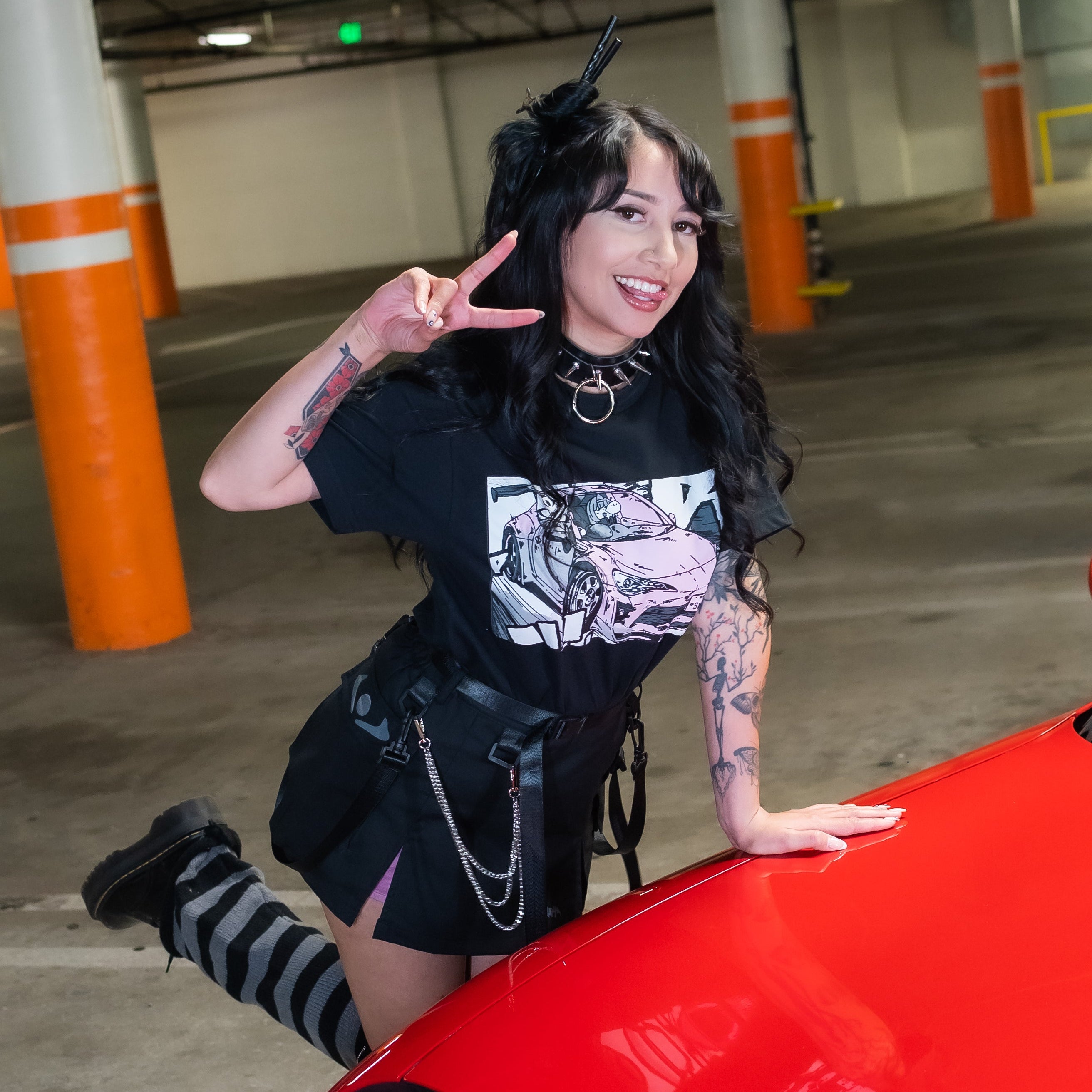 A woman posing in a parking garage wearing a black graphic t-shirt, black shorts, and striped knee-high socks, leaning on a bright red car hood.