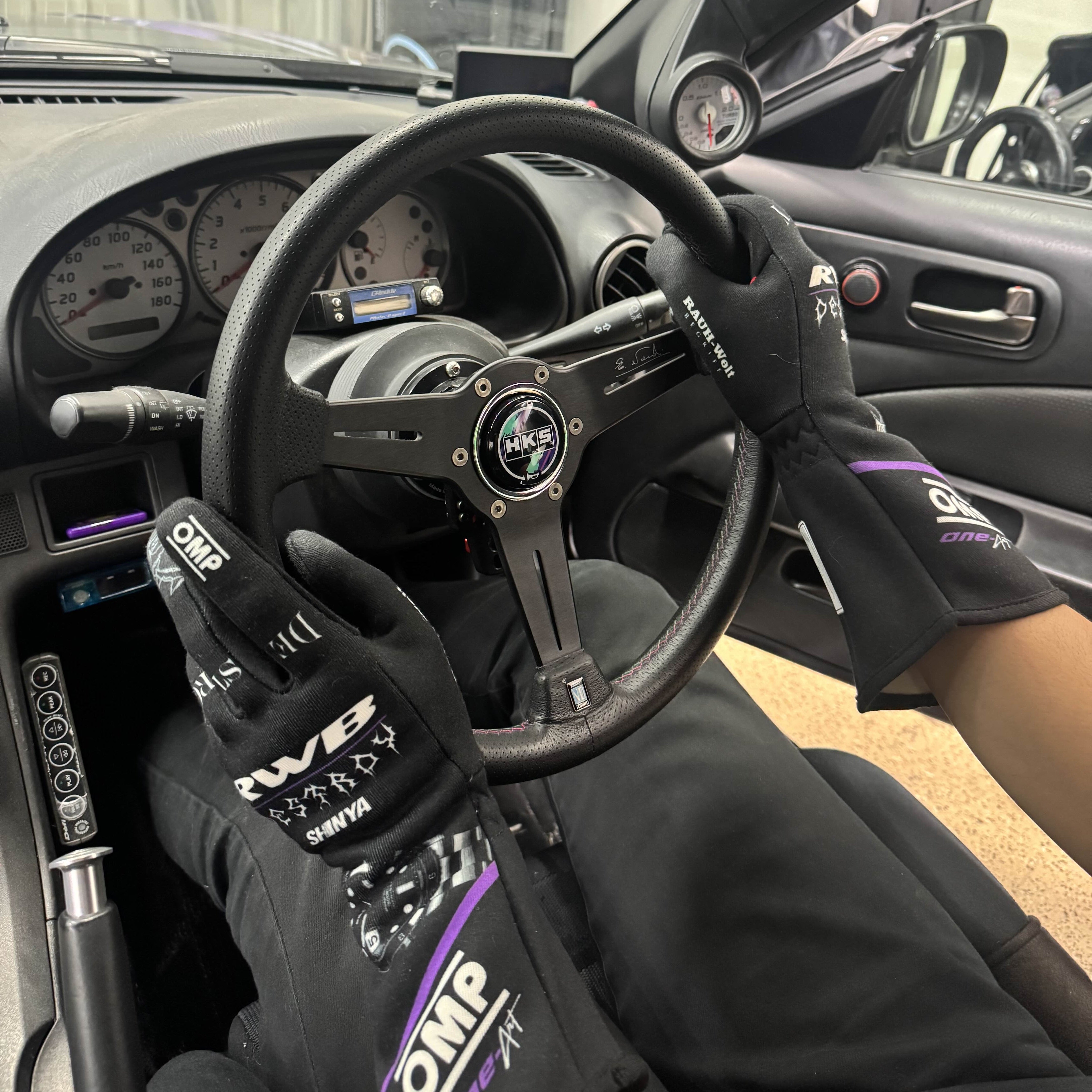 Person wearing gloves with visible branding inside a car, holding a steering wheel.