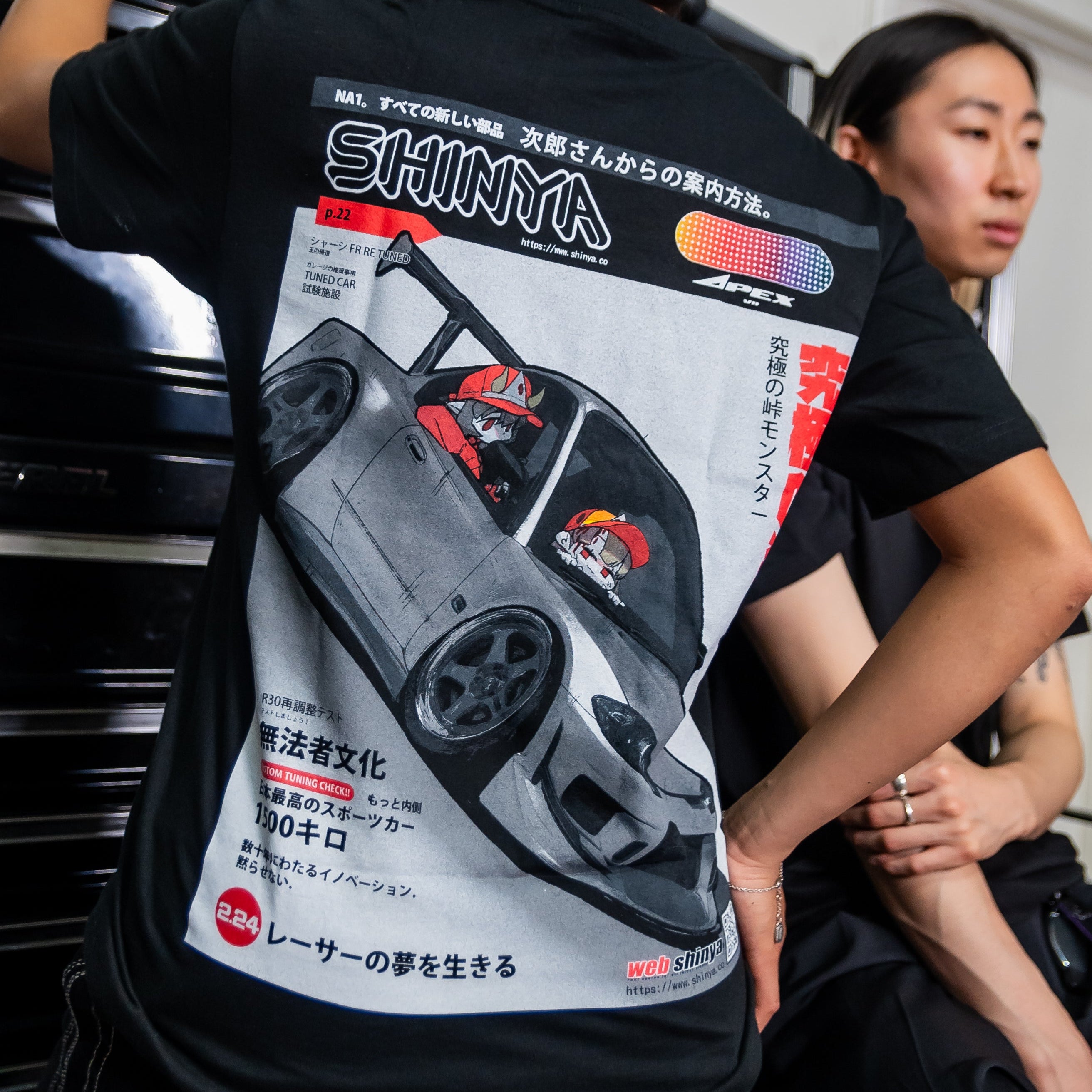 A person wearing a black graphic t-shirt with a large back print of a silver sports car and Japanese text, standing in a garage-like environment next to a tool chest.