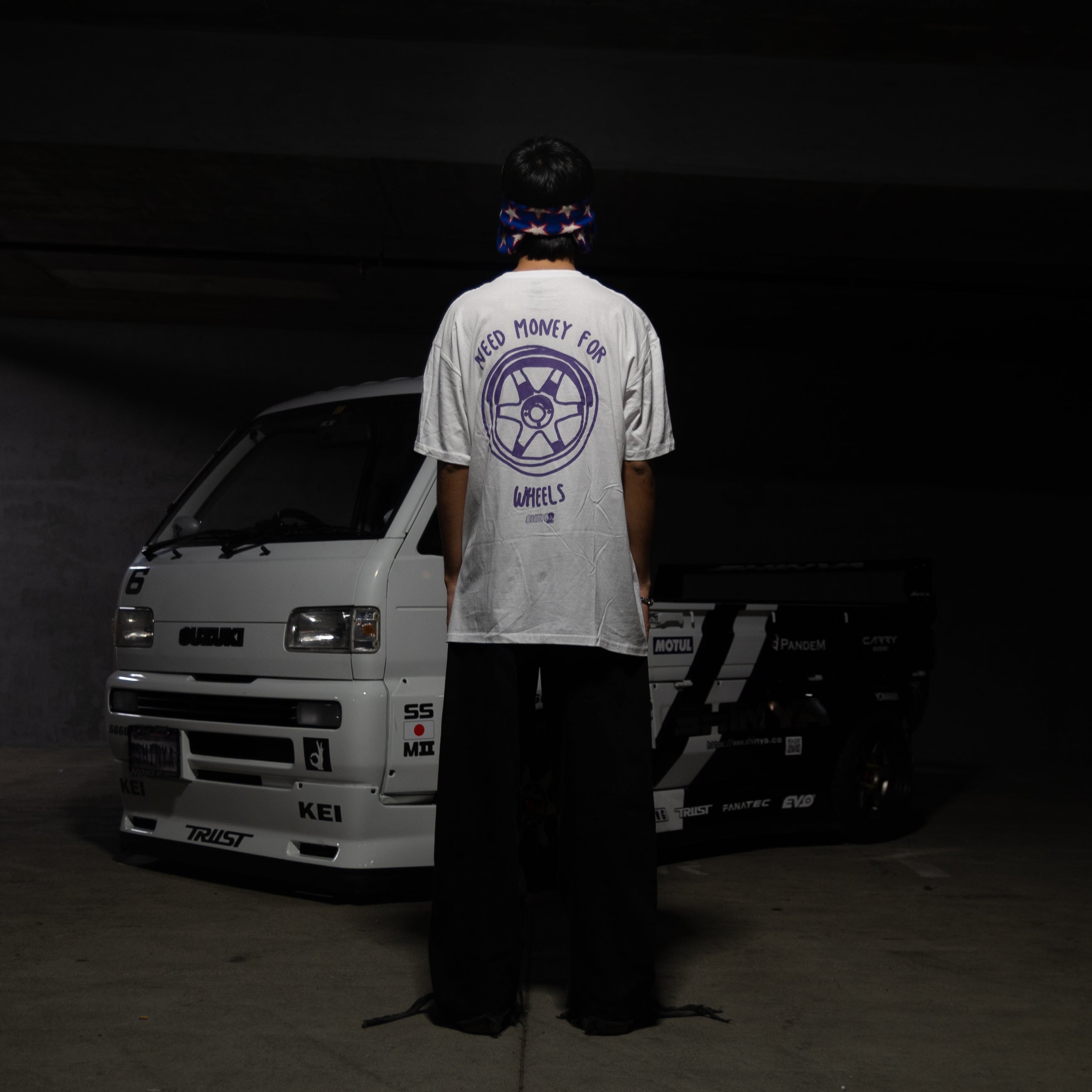 A person stands with their back to the camera in a dark indoor setting, wearing a white graphic t-shirt with a blue circular logo on the back and black pants. A white van is parked behind them.