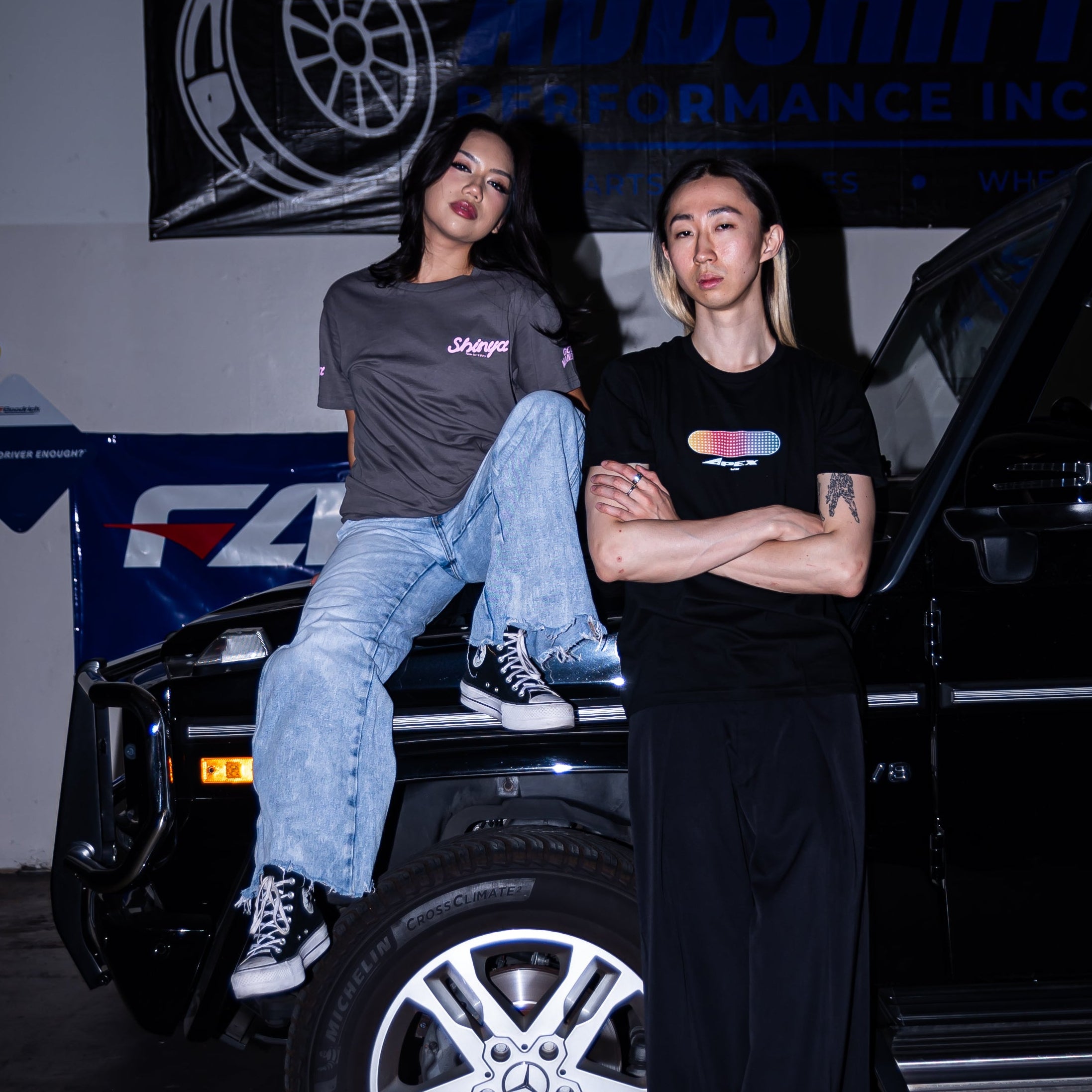 Two people posing with a black vehicle in front of a wall banner that reads 'ADDSHIFT PERFORMANCE INC.' The person on the left sits on the hood wearing a gray t-shirt and light blue jeans, while the person on the right stands with arms crossed wearing a black t-shirt and black pants.