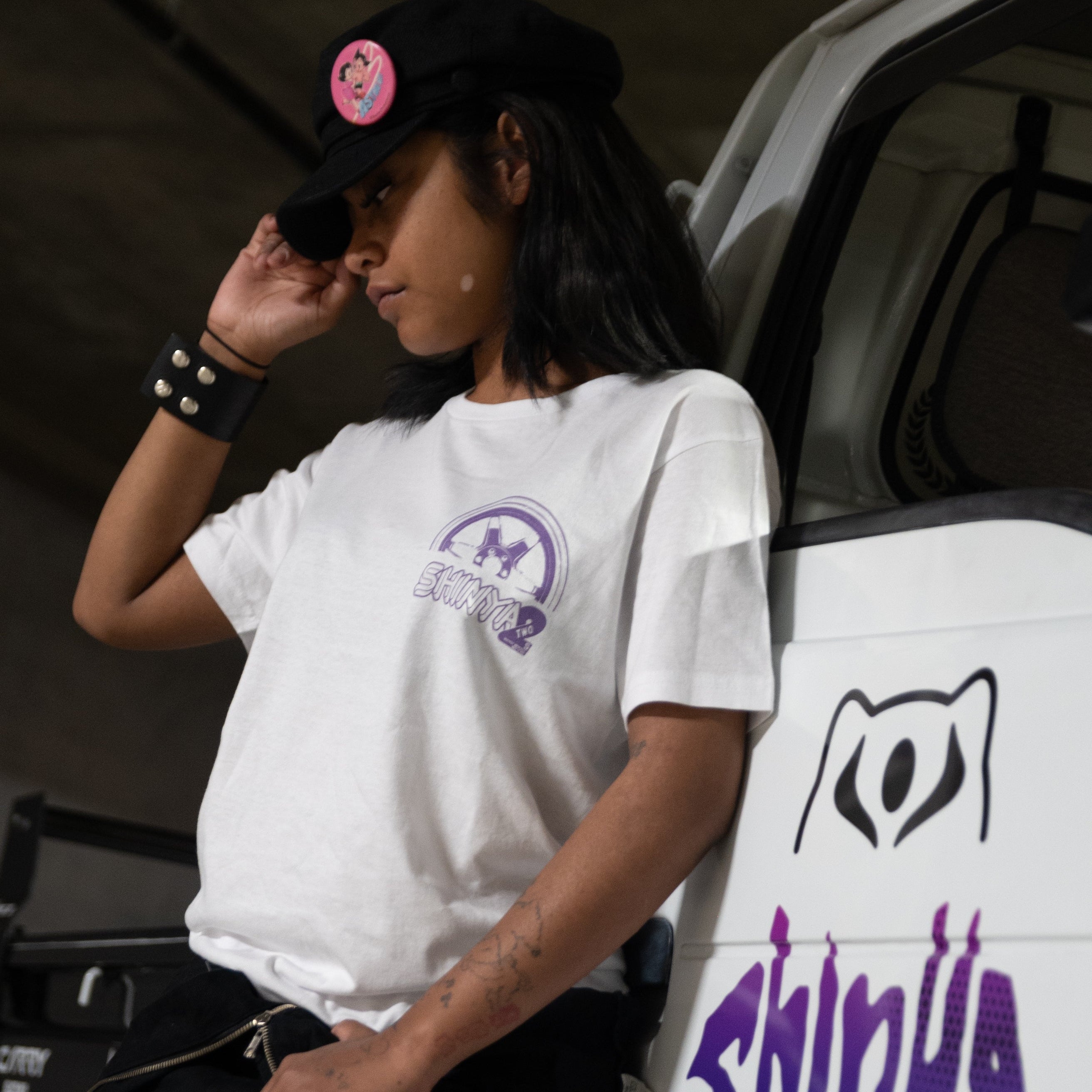 A person wearing a white graphic T-shirt and black cap leans against a white vehicle with purple graffiti-style lettering. The shirt has a small purple chest graphic, and the cap has a pink circular patch.
